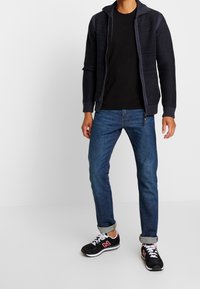 Stickad mörkblå cardigan med dragkedja fram, texturerade ärmar, svart t-shirt under, blå jeans och svarta sneakers med röda detaljer.