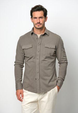 Camicia marrone a maniche lunghe con bottoni, dotata di due tasche sul petto e bottoni a pressione argentati. Tessuto morbido, orlo arrotondato, indossata sopra una maglietta di colore chiaro.