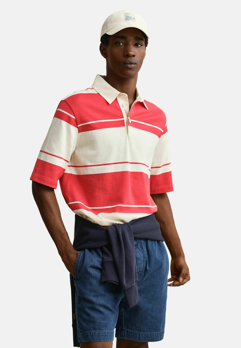 Jeune homme portant un polo rayé rouge et blanc, un short en jean, un sweat-shirt bleu marine noué à la taille et une casquette blanche, debout les mains dans les poches.