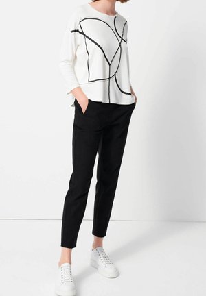 Donna con top bianco a maniche lunghe con linee astratte nere, pantaloni neri affusolati e sneakers con zeppa bianche, in piedi con le mani nelle tasche.