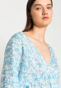 Vestido azul com padrão floral, feito de tecido leve, apresentando um decote em V profundo, mangas longas e uma cintura franzida para um ajuste descontraído.