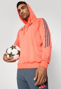 Hettegenser i lys korall med grå striper på ermene. Har kengurulomme og logo-detalj. Holder en mønstret fotball.