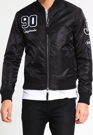 Schwarze Bomberjacke mit durchgehendem Reißverschluss, weißer Text "90 Originals" auf der Brust, zusätzlichem Aufnäher am Ärmel, getragen über einem weiß gesprenkelten Hemd.