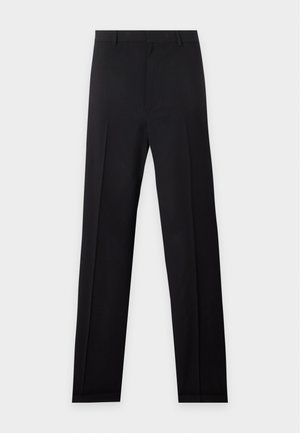 Femme debout avec une main dans la poche, portant un haut noir à manches courtes, un pantalon noir à jambes larges et des sandales noires ouvertes à l'avant.