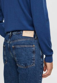 Persona que lleva un suéter azul y unos clásicos jeans de mezclilla azul con una etiqueta beige de Esprit New York en la parte trasera de la cinturilla.