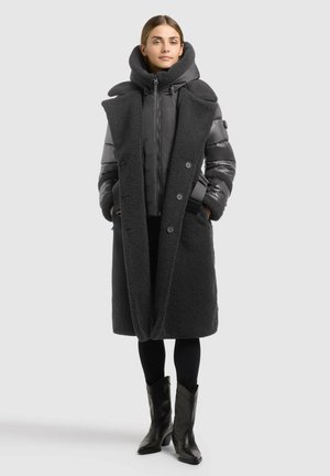 Femme portant un long manteau en shearling noir par-dessus une veste matelassée, des leggings noirs et des bottines noires à talons, debout devant un fond uni.