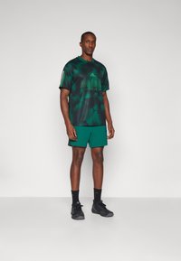 Grønn camo-mønstret treningstrøye med Adidas-logo, kombinert med ensfargede grønne shorts. Brukt med svarte treningssko og sokker.