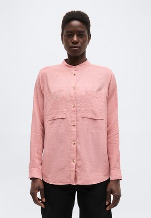 Άτομο που φοράει ένα μακρυμάνικο ροζ πουκάμισο με κουμπιά και δύο μπροστινές τσέπες, στέκεται μπροστά σε ένα απλό ανοιχτό φόντο.