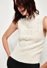 Maglione senza maniche di colore crema con collo alto, caratterizzato da motivi in maglia a trecce testurizzati e accenti decorativi di perle. Tessuto morbido e lavorato a maglia.