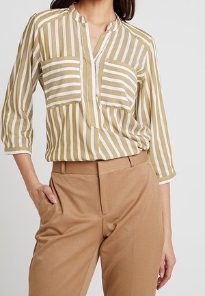 Kvinde iført beige og hvide vertikalt striped bluse med to store vandret striped brystlommer og beige høj talje bukser.