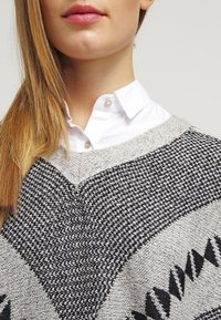 Kobieta z długimi blond włosami, ubrana w białą koszulę z guzikami pod czarnym i szarym swetrem w wzory. 