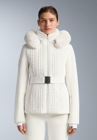 Giacca bianca con trama a maglia intrecciata, dotata di cappuccio con bordo in pelliccia ecologica, chiusura con cerniera e vita con cintura. Maniche lisce completano il modello.