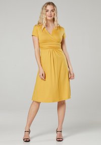 Gelbes Kurzarmkleid mit Wickeldesign, geraffter Taille, knielangem Rock und glatter Textur; ergänzt mit schwarzen, geschnallten Absätzen.
