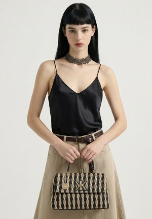 Donna con capelli lisci neri che indossa un camisole di seta nero, gonna beige, cintura marrone, collare ornato e tiene una borsa a motivo.