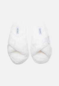 Baltas, pūkains krusta-siksniņu čības, kas izgatavotas no mīksta materiāla, ar strukturētu virsmu un diskrētu logo etiķeti uz iekšējās zoles.