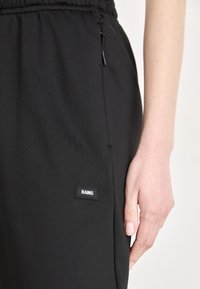 Sorte treningsbukser laget av lett materiale, med elastisk midjebånd, sidelomme med glidelås, og en liten merketag.