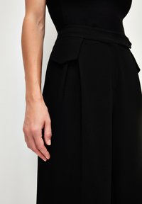Zwarte wijde broek met een hoge taille, voorzien van plooien en een gladde stoffen textuur. Close-up van de tailleband en hand.