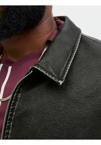 Mustat teksturoidut nahkatakit, joissa on voimakas kaulus, hopeavetoketju ja kerroksellinen viininpunainen paita alla. Sisältää hienovaraisen kiillon.