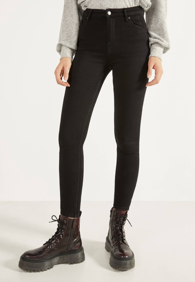 Svarta högmidjade skinny jeans med en slät yta, femfickdesign och knäppning, kombinerade med svarta kängor.