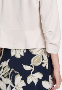 Jasno beżowa, krótka kurtka z rękawami trzy-czwartymi, zestawiona z granatową spódnicą z białymi i zielonymi kwiatowymi wzorami.