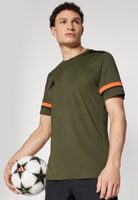 Olijfgroen sportshirt met zwarte accenten en oranje gestreepte mouwen. Lichtgewicht materiaal. Model houdt een met patroon versierde voetbals.