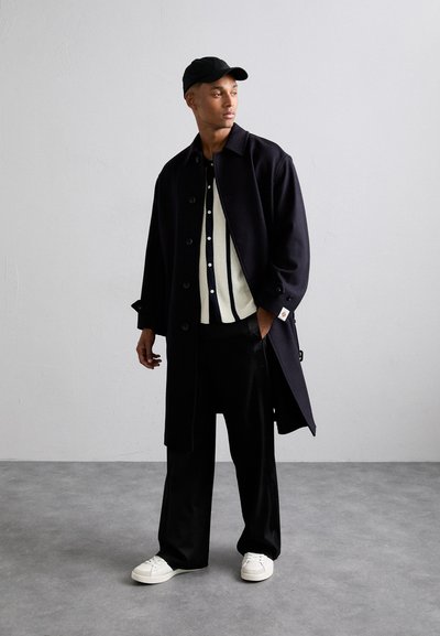 Dlouhý černý kabát s knoflíkovým zapínáním, vrstvený přes bílý žebrovaný svetr s námořnickými pruhy, kombinovaný s černými širokými kalhotami a bílými teniskami.