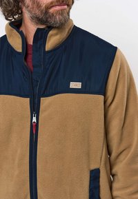 Homme portant une veste polaire marron avec des épaules et un col bleu marine, partiellement zippée, laissant apparaître une chemise bordeaux en dessous.