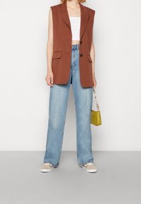 Brun ärmlös kavaj med två fickor fram, tillsammans med lösa ljusblå jeans. Handhållen texturerad gul väska och bruna sneakers.
