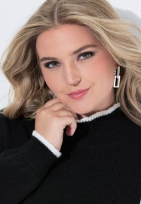 Maglione nero a coste con rifiniture bianche, orecchini d'argento che assomigliano a un anello di catena, texture liscia e onde leggere nei capelli biondi.