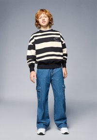 Pulover z črnimi in bež črtami, okroglim izrezom; modri cargo jeans z dvema stranskima žepoma; bele superge; enobarvna siva ozadje.