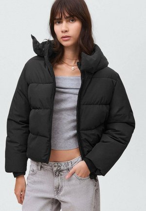 Chaqueta de plumas negra con cuello alto y cierre de cremallera, que presenta un diseño acolchado y un corte relajado. Combinada con una camiseta gris corta y jeans.