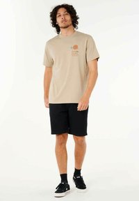 Camiseta beige de manga corta con diseño impreso, combinada con pantalones cortos negros y zapatillas negras. Ajuste simple y relajado, fabricada en algodón.
