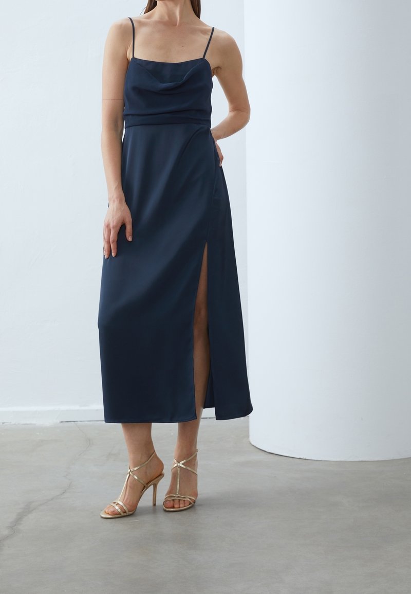 Femme portant une robe bleu marine sans manches avec une fente haute à la cuisse et des sandales dorées à talons hauts et à brides, debout à l'intérieur.