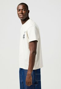 Camiseta de manga corta color crema con cuello redondo, que presenta un pequeño gráfico impreso en el lado izquierdo del pecho y un corte relajado. Pantalones vaqueros visibles.