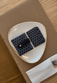 Due portacarte in pelle blu navy con un motivo a onda, dotati di chiusura a scatto, posizionati su un piatto di ceramica bianca. Una forchetta e un tovagliolo sono nelle vicinanze.