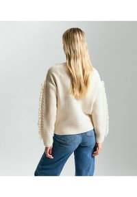 Suéter de punto color crema con acentos de flecos en las mangas, puños acanalados y un ajuste holgado. Llevado con jeans de mezclilla azul.