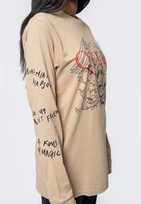 Beige long-sleeve t-shirt with graphic print in red and black. Features winged design and text along the sleeves. Soft, cotton material.