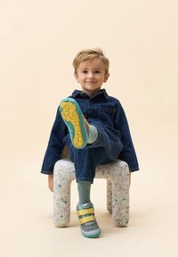 Scarpe sportive grigie e teal con cinghie gialle, abbigliamento in denim, seduto su uno sgabello chiaro macchiato contro uno sfondo beige.