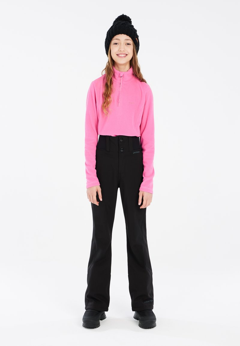 Roze fleece pullover met halve rits, gecombineerd met hoge zwarte broeken met twee knopen. Een zwarte gebreide muts en stevige zwarte schoenen maken de look compleet.