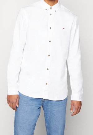 Man wearing fitted white long-sleeve button-up shirt with small logo on chest and light blue jeans against plain background.
