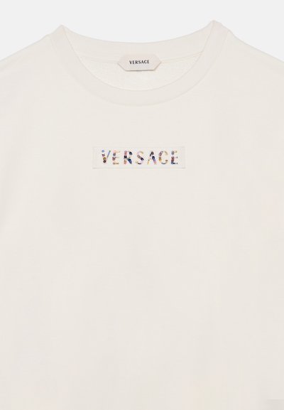 T-shirt Versace couleur crème avec un col rond et le nom de la marque écrit en petites perles colorées sur un patch rectangulaire sur la poitrine.