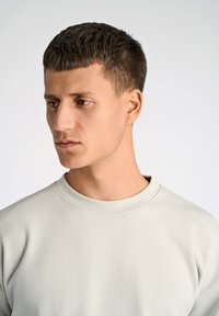 Sudadera gris claro con cuello redondo, textura suave y detalles de costura mínimos, llevada por una persona que está mirando ligeramente hacia la derecha.