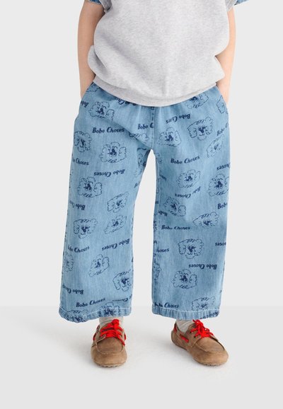 Enfant portant un pantalon ample en jean bleu clair avec des motifs de nuages et "Bobo Choses", des chaussures beiges avec des lacets rouge vif, et un haut gris.