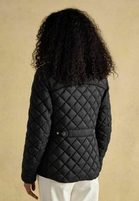 Čierna prešívaná bunda s priliehavým strihom, zdobená diamantovými vzormi, detailom v podobe opasku na chrbte a patentkami na páse.