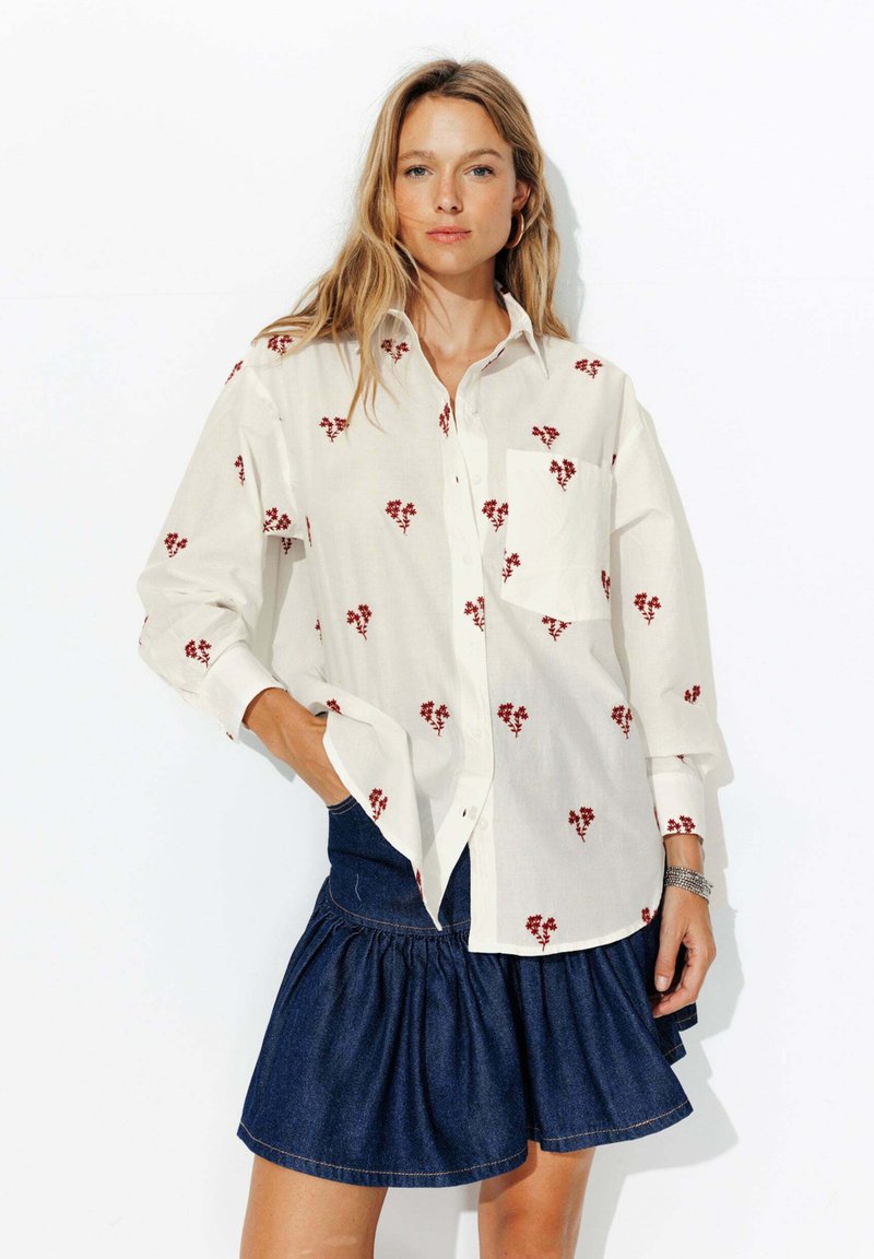 Chemise blanche à boutons avec broderie florale rouge, manches longues et poche avant, associée à une jupe en denim avec tissu froncé.
