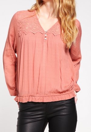 Mujer que lleva una blusa coral suelta de manga larga con canesú de encaje y botones en el escote, combinada con pantalones de cuero negro.