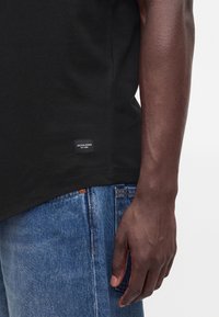 Άτομο που φοράει μαύρο μπλουζάκι Jack & Jones και μπλε τζιν, εμφανίζεται από τον ώμο έως τον μηρό, με το δεξί χέρι χαλαρό δίπλα στο σώμα.