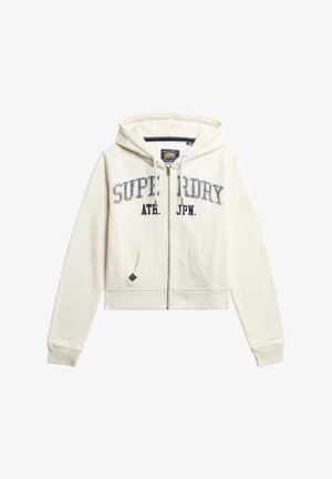 Sudadera corta color crema con cremallera frontal, que presenta bordado en texto "SUPERDRY ATH. JPN" en azul marino, dos bolsillos frontales y puños acanalados.