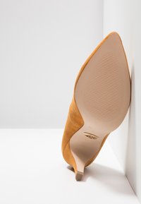 Semišové vysoké podpatky v hořčicově žluté barvě, s špičatou špičkou a úzkým stiletto podpatkem. Hladká podrážka s vroubkovanou texturou a kovovým logem.