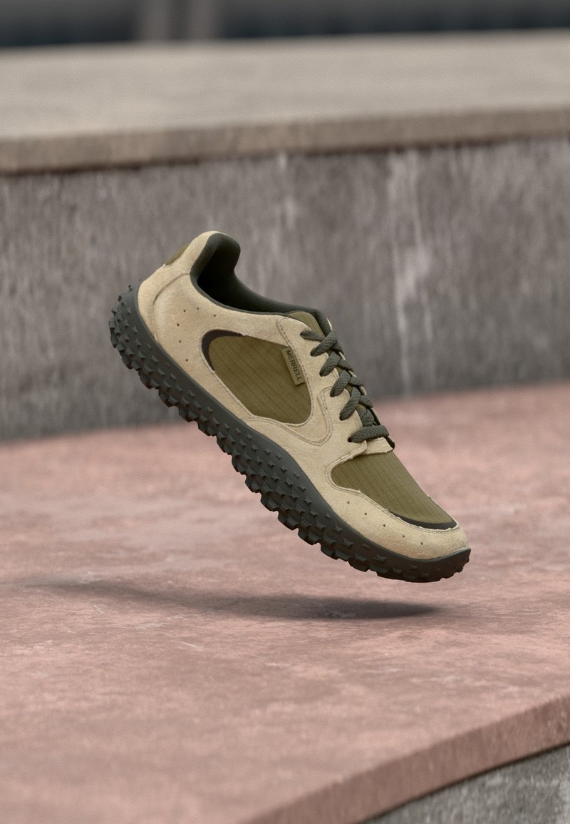 Ανοιχτό καφέ και ελιά πράσινο παπούτσι πεζοπορίας με υφή μαύρης σόλας αιωρούμενο στον αέρα πάνω από επιφάνεια από πέτρα.